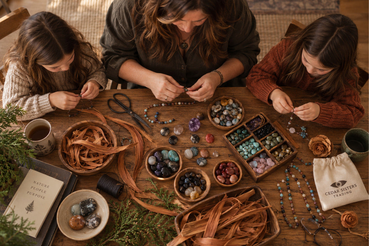 Three women sit around a rustic wooden table crafting with beads, leather strips, and crystals in a warm, cozy setting. A small white drawstring pouch on the table is branded with 'CROW SKILLED CRAFTING'.