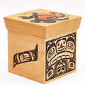 A square wooden box with a hinged lid, made from light natural wood, decorated with black and red Indigenous-style totem and face carvings on the lid, front, and side.