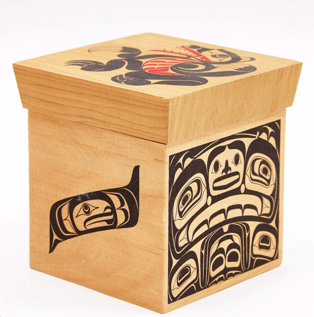 A square wooden box with a hinged lid, made from light natural wood, decorated with black and red Indigenous-style totem and face carvings on the lid, front, and side.